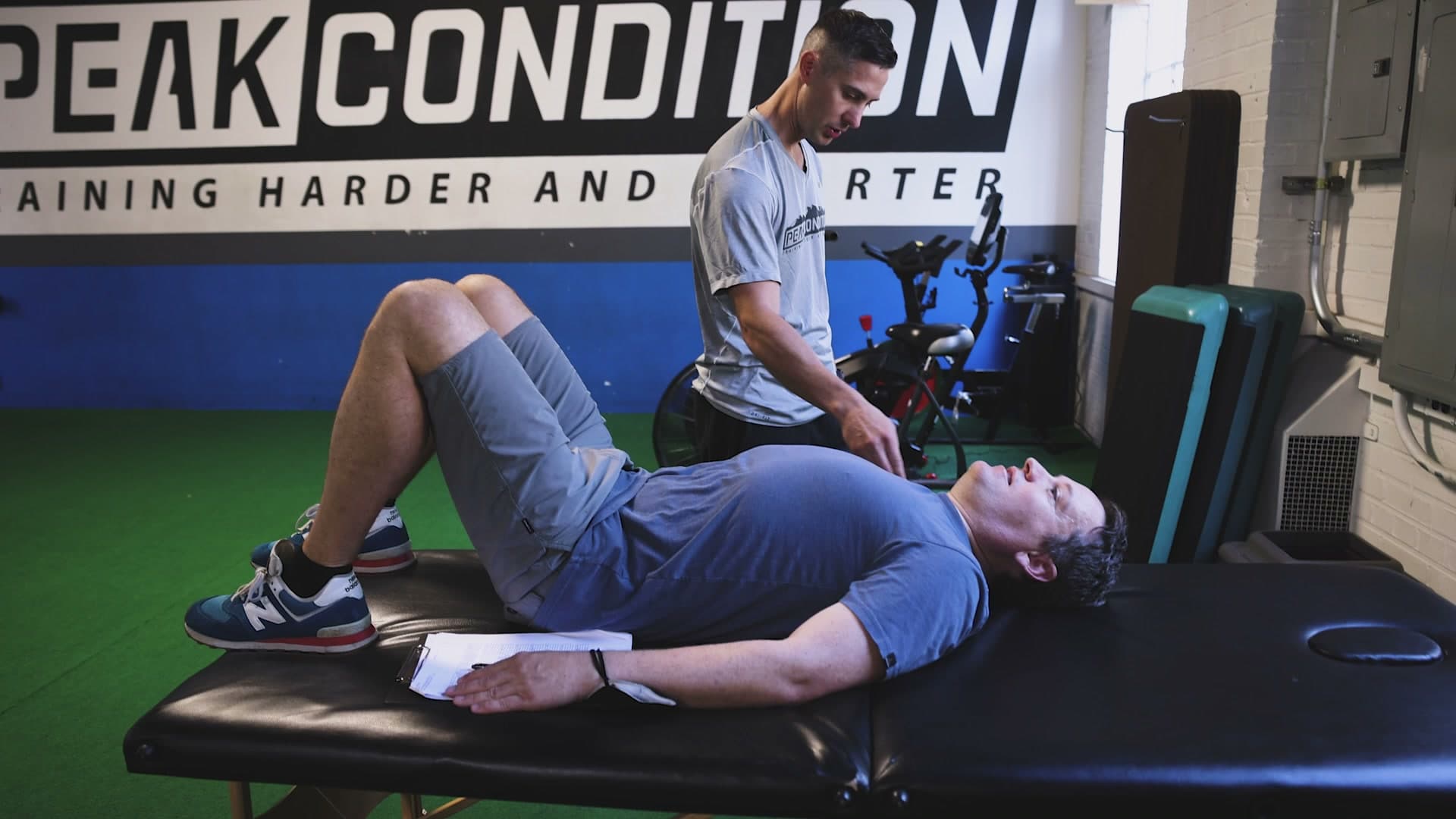 A coach working through a movement with an athlete on a bench, clipboard in hand